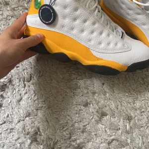 Nike Jordans 13 - Säljer ett par snygga Nike Air Jordans i vitt och gult. Skorna har en cool design med svart sula och röda detaljer. Perfekta för både vardag och sportiga aktiviteter. De är i nytt skick och har aldrig använts. Passa på att köpa dessa unika sneakers! 