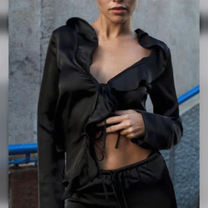 Svart satinblus med volangdetaljer - Snygg svart blus i satin med volangdetaljer och knytning framtill. Perfekt för en kväll ute eller en fest. Den har långa ärmar och en elegant, glansig finish som ger en lyxig känsla. Storlek M men passar ej mig som bär M utan mer någon som har S