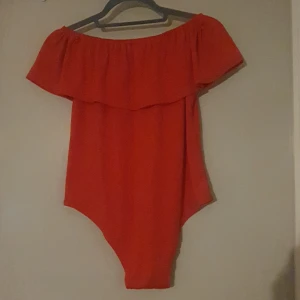 Röd body med volang - Säljer en superfin röd body med volangdetaljer. Perfekt för sommaren eller under en kofta.. Den har en off-shoulder design som ger en trendig look. Materialet känns mjukt och bekvämt mot huden. 