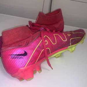 Säljer ett par Nike Mercurial fotbollsskor i en cool rosa färg med gula detaljer. Skorna har snörning och är i syntetmaterial, perfekt för fotbollsträning. De har en tight passform och är i mycket bra skick. Sulan är designad för optimalt grepp på planen.