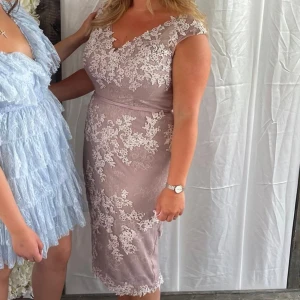 Beige spetsklänning - Elegant beige klänning med vackra spetsdetaljer. Den har en v-ringad hals och är ärmlös, vilket ger en sofistikerad look. Perfekt för sommarens fester eller bröllop. Klänningen är knälång och har en figurnära passform som framhäver silhuetten.