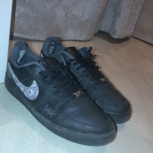 Svarta Nike Air Force 1 med custom design - Säljer ett par svarta Nike Air Force 1 med en unik custom design på swooshen. Skorna har snörning och är i acceptabelt skick med synligt slitage på insidan. Perfekta för dig som vill ha en personlig touch på dina sneakers!