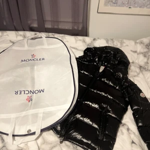 Svart pufferjacka från Moncler - Säljer en sprillans ny snygg svart pufferjacka från Moncler. Jackan har en glansig finish och är perfekt för kalla dagar. Den har en dragkedja framtill och en huva för extra värme. Moncler-loggan syns på ärmen. Perfekt för vintern! ❄️P.S SÅ KLART DEN BLIR BLIGGARE VID SNABB AFFÄR