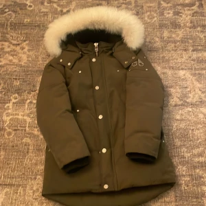 Olivgrön dunjacka från Moose Knuckles - Säljer en olivgrön dunjacka från Moose Knuckles i mycket bra skick. Jackan har en fluffig pälsfodrad huva och stängs med dragkedja och knappar. Perfekt för kalla vinterdagar! 🧥❄️