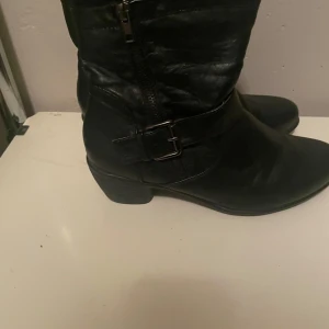 Svarta boots med spänne - Snygga svarta boots i skinn med dragkedja och spänne på sidan. Perfekta för höst och vinter med en robust design och låg klack. De har en klassisk look som passar till både vardag och fest. Använd dem för att ge din outfit en cool touch!