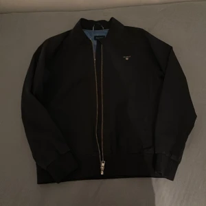 Svart bomberjacka från GANT - Säljer en snygg svart bomberjacka från GANT i storlek 146/152 cm, perfekt för 11-12 åringar. Jackan har en dragkedja framtill och är broderad med GANT-loggan på bröstet. Insidan är fodrad i blått med ett stort GANT-tryck. Passar perfekt för höst och vår!