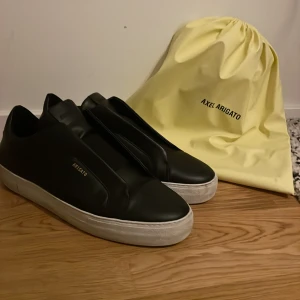 Svarta sneakers från Axel Arigato - Säljer ett par stilrena svarta sneakers från Axel Arigato. De är i mycket bra skick och kommer med originalpåse. Perfekta för både vardag och fest med sin enkla men eleganta design. Skorna har en vit sula som ger en snygg kontrast till det svarta.