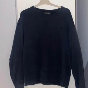 Svart tröja från Acne Studios - Säljer en svart tröja från Acne Studios med deras ikoniska ansiktsmärke på bröstet. Tröjan har långa ärmar och en liten reva på ena ärmen. Tröjan har även några vita prickar/fläckar. Går nog bort i tvätten. 