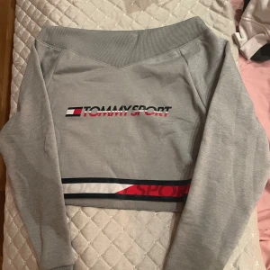 Grå croppad tröja från Tommy Sport - Säljer en snygg grå croppad tröja från Tommy Sport. Den har en cool logga på framsidan och en bred ribbad kant som gör den of sholder. Perfekt för en sportig look eller en avslappnad dag. Passar bra till jeans eller träningsbyxor!