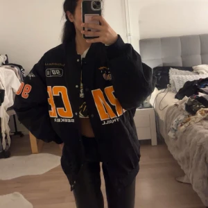 Svart bomberjacka med tryck - Säljer en svart bomberjacka med coola tryck i orange och vitt. Jackan har knappar framtill och är i oversized passform. Perfekt för höst och vår, och ger en sportig look. Passar både till vardags och fest!
