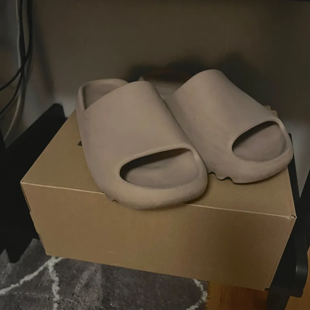 Säljer ett par bekväma och stilrena beige yeezy slides från Adidas. De har en ribbad sula för extra grepp och komfort. Passar både till stranden och vardagsbruk. Använda men i bra skick!. Skor.