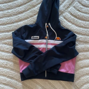 Svart och rosa hoodie från Ellesse - Säljer en snygg och bekväm hoodie från Ellesse i svart och rosa. Den har en dragkedja framtill och en justerbar huva med vita snören. Perfekt för en avslappnad stil eller träning. Den är croppad och har ribbade muddar vid ärmslut och nederkant💗