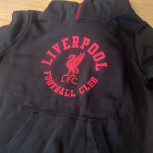 Säljer en svart hoodie från Liverpool FC med röd logga och text på framsidan. Den har en mysig huva och en stor ficka framtill. Perfekt för fotbollsfans som vill visa sitt stöd för laget. Passar bra till både vardag och matchdagar! Aldrig använd men har råkat ta av prislappen