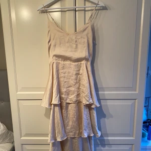 Champagnefärgad klänning med volanger - Säljer en elegant champagnefärgad klänning med volanger. Den har smala axelband och en flödande design som ger en romantisk känsla. Perfekt för sommarfester eller bröllop. Klänningen är i mycket bra skick och har en vacker, lätt skimrande yta.