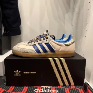 Snygga Adidas Wales Bonner sneakers i beige och blått. De har en cool retrostil med blå ränder och mönstrade snören. Skorna är i skinn och mocka med en gummisula som ger bra grepp. Perfekta för både vardag och fest!