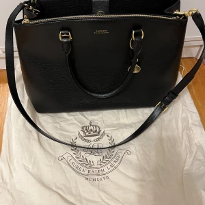 Svart axelväska från Lauren Ralph Lauren - Förhandla gärna!!Snygg svart axelväska i skinn från Lauren Ralph Lauren. Väskan har guldfärgade detaljer och en praktisk dragkedja upptill. Perfekt för både vardag. Den är rymlig och har en stilren design som passar till allt.