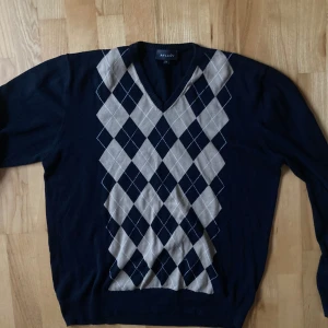 Mörkblå argyle-mönstrad tröja från Avalon - Säljer en stilren mörkblå tröja från Avalon med klassiskt argyle-mönster i ljusgrått och vitt. Tröjan har en v-ringad hals och långa ärmar, 