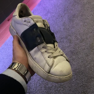 Valentino sneakers i vitt och mörkblått - Säljer ett par Valentino sneakers i vitt och mörkblått. Skorna har en cool design med en bred svart rem över mitten. De är i bra skick men har några tecken på användning. Perfekta för en stilren och avslappnad look! Box finns