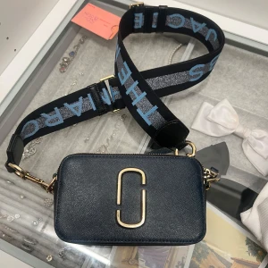 Marinblå axelväska från Marc Jacobs - Snygg marinblå axelväska från Marc Jacobs med ett stort guldspänne framtill. Väskan har en bred, justerbar axelrem med text i blått och silver. Den är perfekt för både vardag och fest. Två dragkedjor upptill ger enkel åtkomst till dina saker.