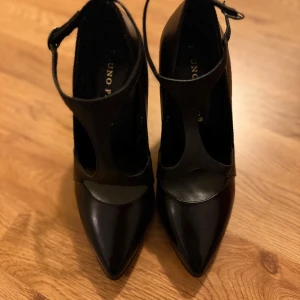 Svarta klackskor från Uno Piu - Snygga svarta klackskor från Uno Piu med spetsig tå och elegant design. Skorna har en stilren rem över foten och en bekväm klack som ger extra höjd. Perfekta för festliga tillfällen eller en utekväll. De är i mycket bra skick och redo att användas!