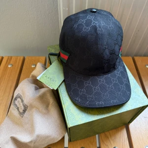 Gucci keps - Säljer en snygg svart keps från Gucci med deras ikoniska mönster. Kepsen har en justerbar rem baktill och detaljer i rött och grönt på sidan. Perfekt för att ge din outfit en lyxig touch. Kommer med originalförpackning och dustbag. Obs:Inga returer