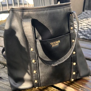Fin väska från Victoria's Secret! - Fin väska i skinnimmitation med många fack och dekorativa nitar. Den har både handtag och justerbar axelrem. Mått: 35 x 35 cm. Små spår av användning finns men den är fortfarande i mycket gott skick. 