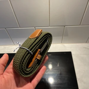 Flätat bälte - Säljer ett snyggt olivgrönt flätat bälte med stilrena bruna läderdetaljer. Perfekt för att ge en touch av stil till vilken outfit som helst. Bältet har ett silverfärgat spänne som ger en klassisk look. Passar både till vardags och festliga tillfällen. Bältet är väldigt elastiskt och passar alla storlekar! 