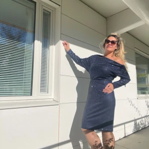 Glittrig mörkblå offshoulder klänning - Snygg glittrig mörkblå klänning med offshoulder-design. Perfekt för festliga tillfällen! Klänningen har en tight passform som framhäver figuren och är tillverkad i ett bekvämt material. Den är knälång och har långa ärmar, vilket gör den idealisk för både höst och vinter.