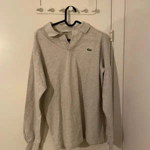 Grå långärmad pikétröja från Lacoste - Säljer en klassisk grå långärmad pikétröja från Lacoste. Den har en stilren design med krage och knappar framtill. Perfekt för både vardag och lite finare tillfällen. Tröjan är i bra skick och redo för en ny ägare!
