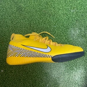 Gula Nike Mercurial fotbollsskor - Säljer ett par coola gula Nike Mercurial fotbollsskor i bra skick. De har en snygg vit Nike-logga på sidan och ett häftigt mönster i svart och vitt runt hälen. Perfekta för inomhusfotboll eller konstgräs. Skorna har snörning och en tight passform för bästa kontroll på planen.