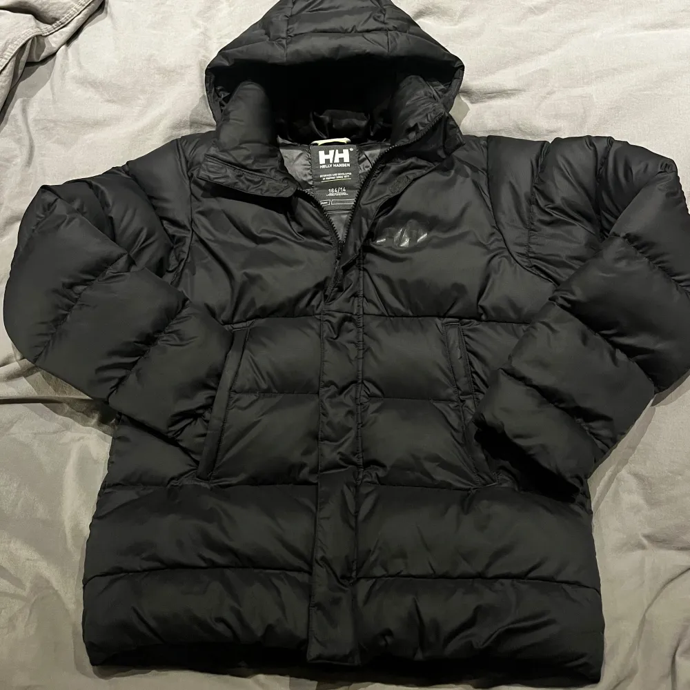 Säljer en svart dunjacka från Helly Hansen i storlek 164. Jackan är perfekt för kalla vinterdagar med sin tjocka vaddering och huva. Den har en stilren design med Helly Hansen-logga på bröstet och ryggen. Jackan är i mycket bra skick och redo för nya äventyr! 🖤Luvan är avtagbar. . Takit.