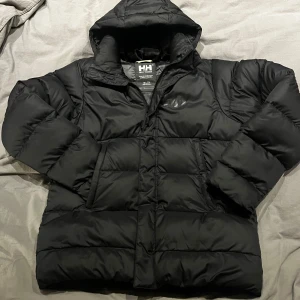 Svart dunjacka från Helly Hansen - Säljer en svart dunjacka från Helly Hansen i storlek 164. Jackan är perfekt för kalla vinterdagar med sin tjocka vaddering och huva. Den har en stilren design med Helly Hansen-logga på bröstet och ryggen. Jackan är i mycket bra skick och redo för nya äventyr! 🖤Luvan är avtagbar. 