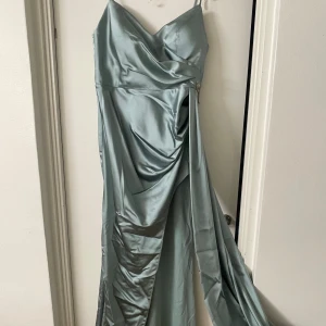 Ljusgrön/blå långklänning med smyckesdetalj - Elegant ljusgrön/blå långklänning i satin med smala axelband och v-ringad hals. Klänningen har en vacker drapering och en glittrig smyckesdetalj i form av en fjäril vid midjan. Perfekt för festliga tillfällen! använd endast en gång i några få timmar och är i perfekt skick , inga defekter