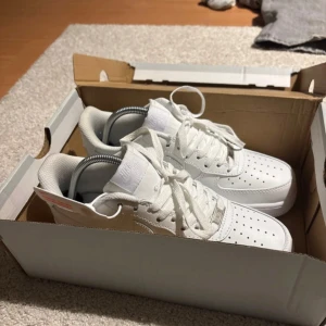 Vita Nike Air Force sneakers - Säljer ett par klassiska vita Nike Air Force sneakers i nytt skick. De är helt vita med snörning och har den ikoniska Nike-loggan på sidan. Perfekta för både vardag och fest, och de passar till nästan alla outfits. Skorna är i storlek herr 42 och kommer i originalkartong.