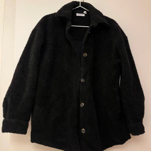 Svart teddyjacka från Weekday - Mysig svart teddyjacka från Weekday i storlek XS, oversize. Perfekt för kyliga dagar med sin fluffiga och varma design. Jackan har knappar framtill och långa ärmar. Ett måste för höst- och vintergarderoben!