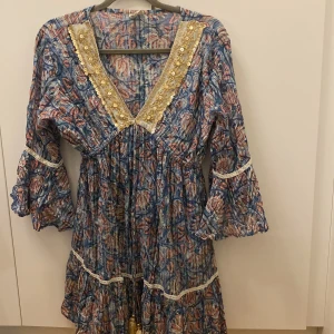 Zimmermann look alike med gulddetaljer - Säljer en superfin blommig klänning i blått och rosa med v-ringad spetskant och gulddetaljer. Den har trekvartsärmar med volang och en lös passform. Perfekt för sommarens fester eller en dag på stan. Klänningen har också dekorativa tofsar framtill. 🌸✨