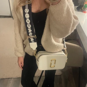 Vit axelväska från Marc Jacobs - Säljer en snygg vit axelväska från Marc Jacobs med guldiga detaljer och en bred, justerbar axelrem. Väskan har en praktisk dragkedja och en stilren design med logotypen på framsidan. Väskan är använd och har fått fåtal defekter, men inget som syns på långt håll eller vid användning 💕😊