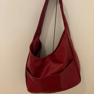 Röd axelväska i syntetmaterial - Snygg röd axelväska i syntetmaterial med en enkel och stilren design. Väskan har en praktisk storlek med en bred axelrem och en knappstängning upptill. Perfekt för vardagsbruk eller en utekväll. Den har en slät yta och en liten ficka på framsidan för extra förvaring. Mycket bra skick och säljer för 150kr 