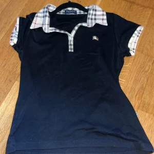 Svart kortärmad topp från Burberry - Säljer en snygg svart topp från Burberry med klassisk rutig krage och ärmslut. Den har en liten broderad logga på bröstet. Perfekt för både vardag och lite finare tillfällen. Passar bra till jeans eller kjol!