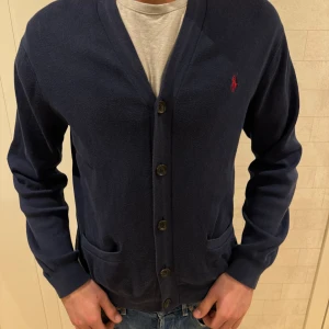 Mörkblå cardigan från Ralph Lauren - Säljer en snygg mörkblå kofta från Ralph Lauren. Den har en klassisk design med knappar framtill och två praktiska fickor. Perfekt för både höst och vår. Den röda loggan på bröstet ger en stilren touch. Passar bra till både jeans och chinos!