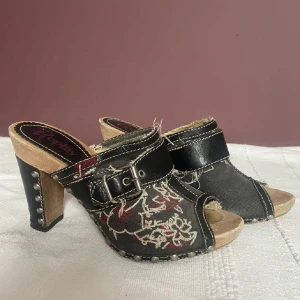 Svarta klackskor med broderi från Replay - Snygga svarta klackskor från Replay med coolt blommigt broderi i rött och vitt. De har en öppen tå och en rejäl klack med nitar på sidan. Skorna är i jeansmaterial med läderdetaljer och ett bälte över foten. Perfekta för en utekväll eller fest!