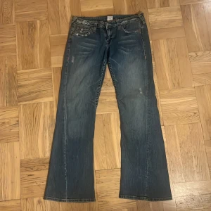 Blå jeans från True Religion - Säljer ett par snygga blå jeans från True Religion i bra skick. De har en bootcut-stil och är dekorerade med coola sömmar och knappar på bakfickorna. Perfekta för en avslappnad look eller en utekväll. Passar till det mesta och är superbekväma!