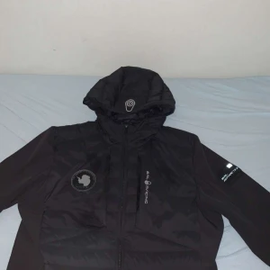 Svart pufferjacka från Sail Racing - Säljer en svart pufferjacka från Sail Racing i storlek medium. Jackan har en cool design med en broderad logga på bröstet och en praktisk huva. Perfekt för kalla dagar med sin quiltade stil som håller värmen. Den har en dragkedja framtill och är långärmad. Passar perfekt för både höst och vinter!