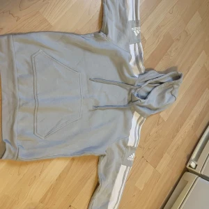 Grå hoodie från Adidas - Säljer en snygg och bekväm grå/vit Hoodie Märke Adidas. Den har de klassiska tre ränderna längs ärmarna och en stor magficka framtill. Perfekt för en avslappnad stil eller träning.                  Tillverkad i Turkiet med Primegreen-material. 