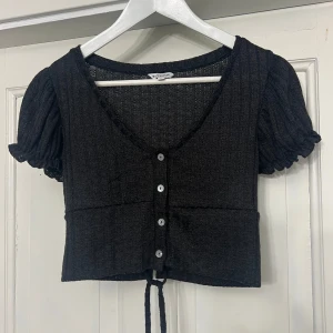 Svart kortärmad topp med knappar och snörning - Säljer min svarta topp med korta puffärmar från Clockhouse. Toppen har knappar framtill och snörning i ryggen. Storlek M och är i nyskick. 
