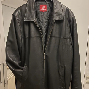 Svart skinnjacka från Redwood - Säljer en snygg svart skinnjacka från Redwood i storlek L. Jackan har en klassisk design med dragkedja och quiltat foder för extra värme. Perfekt för höst och vår! Den har långa ärmar och en stilren look som passar till det mesta. Inuti finns praktiska fickor för förvaring.