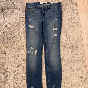 Blå jeans från Hollister - Säljer ett par snygga blå jeans från Hollister i modellen 'Low Rise Super Skinny'. De har en cool slitning på flera ställen som ger en trendig look. Perfekta för en casual outfit eller en utekväll. Storlek W26 L28.