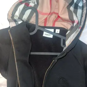 Säljer en snygg svart hoodie från Burberry med klassiskt rutigt mönster i luvan. Den har en dragkedja framtill och snörning vid luvan. Se sista bild för en defekt i fickan, går enkelt att lösa med och sy, storlek S