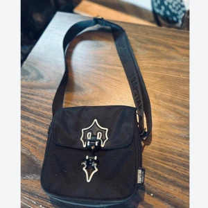 Svart axelväska med spänne - Säljer en snygg svart Trapstar axelväska med snygga detaljer. Väskan har ett unikt spänne framtill och en justerbar axelrem. Den är i nyskick och redo för nya äventyr!