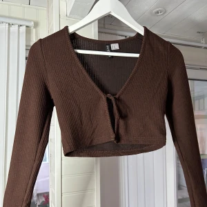 Brun ribbad topp från Gina Tricot - Säljer en snygg brun ribbad topp från H&M. Den är croppad och har en v-ringning med knyt framtill.Superfin att matcha med jeans eller kjol! Samt ta på den över klänning🥰